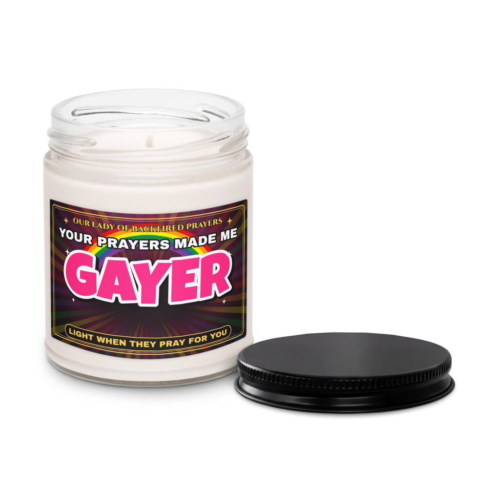Nice Gay soy candle with the lid off beside its black lid, showing white soy wax and a label reading "Our Lady of Backfired Prayers" and "Your Prayers Made Me GAYER."