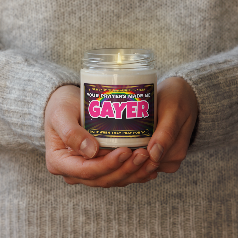 Hands in a gray knit sweater holding the lit Nice Gay soy candle with the lid off, showing its label reading "Our Lady of Backfired Prayers," "Your Prayers Made Me GAYER," and "Light when they pray for you" over a rainbow.