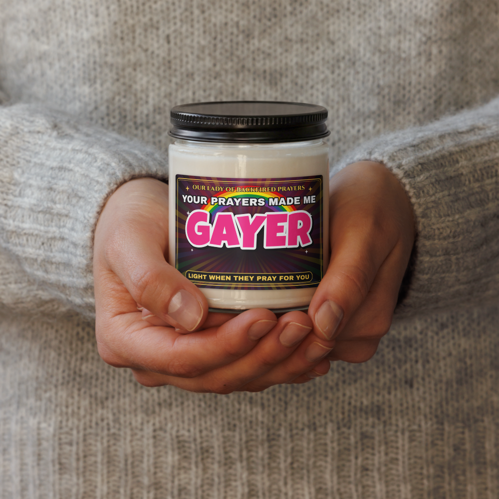 Hands in a gray knit sweater holding the Nice Gay soy candle, lid on, showing its label reading "Our Lady of Backfired Prayers," "Your Prayers Made Me GAYER," and "Light when they pray for you" over a rainbow.